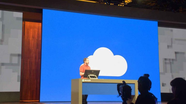 Photo showing a Microsoft employee speaking in front of a large blue screen showing a white cloud OneDrive icon